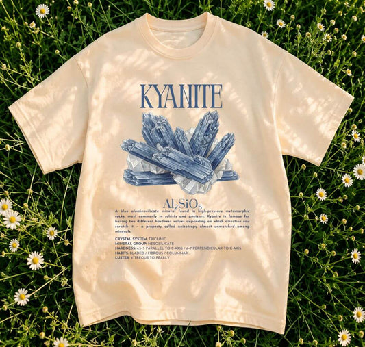 A cream-colored short-sleeve t-shirt with a blue graphic of a crystal cluster and the text 'KYANITE, laid flat on a background of green grass and small white daisies.